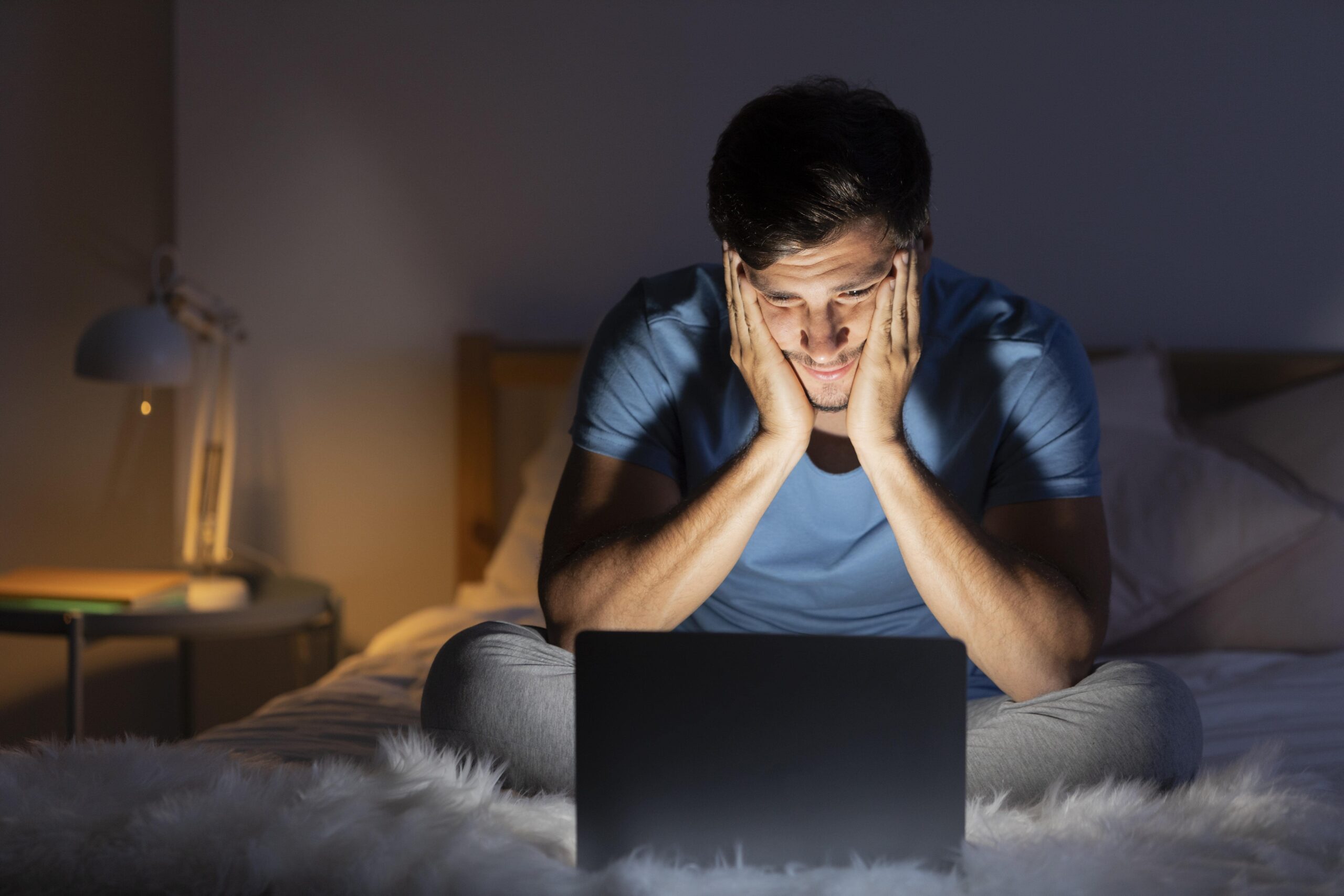 Man sitting on bed, staring at laptop screen late at night, looking stressed