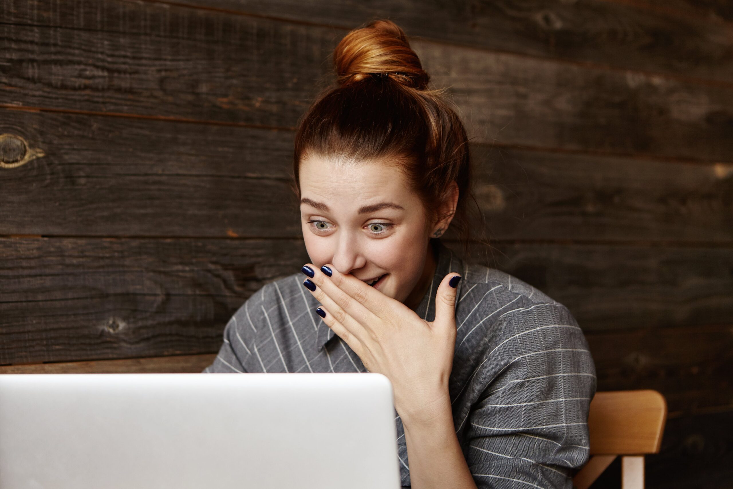 Young woman with a top bun smiling in surprise while looking at a laptop screen, sitting in front of a rustic wooden wall background.